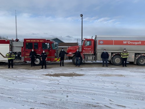 Shows a shot of 7 people outdoors winter with a firetruck and a McDougall fuel truck behind them
