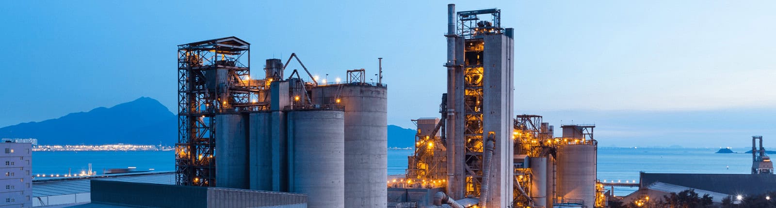 Refinery with tanks and industrial equipment lit up at dusk near water and mountains.