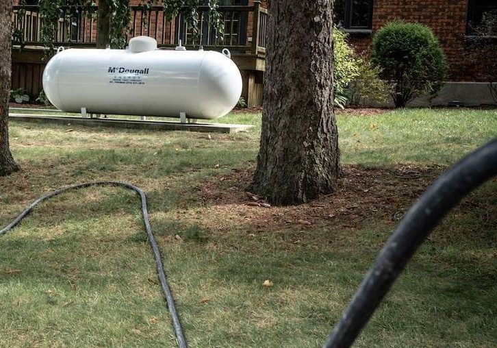 White propane tank installed outdoors on a grassy area near trees and a brick house.