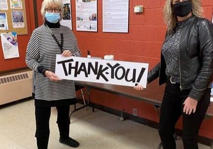 Shows 2 women standing indoors with face masks holding a sign that says Thank You