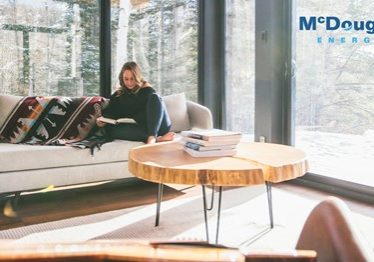 Shows a woman reading a book sitting on a large couch in a large room surrounded by windows