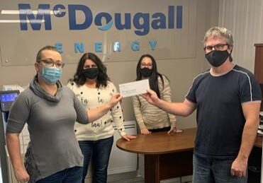 Shows a group of 4 people standing and holding a cheque wearing face masks against wall with McDougall Energy logo