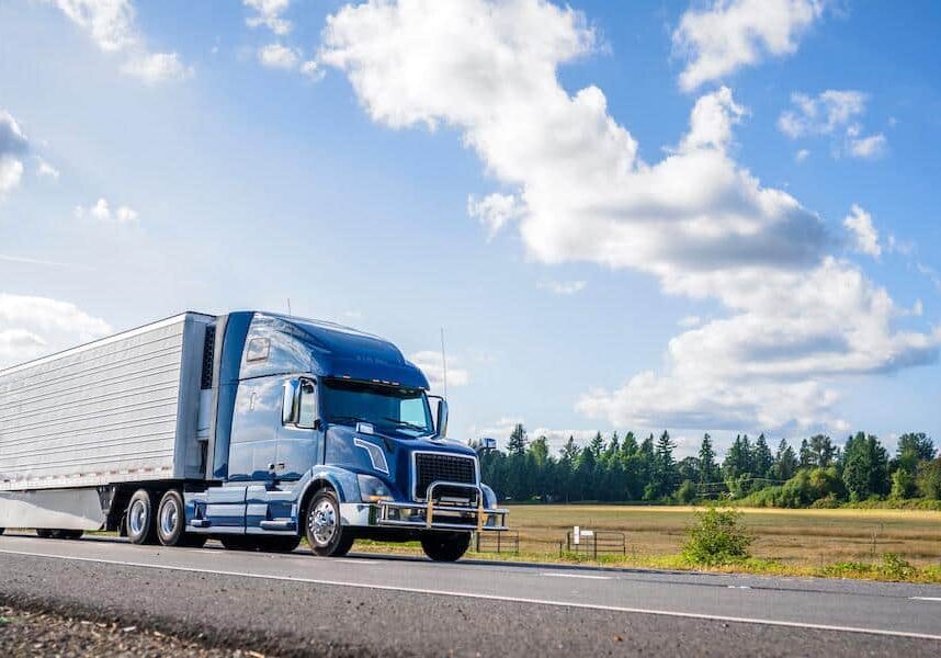 Big rig powerful professional industrial blue bonnet semi truck for long haul delivery commercial cargo going with refrigerator semi trailer on the summer road with forest and meadows on the sides
