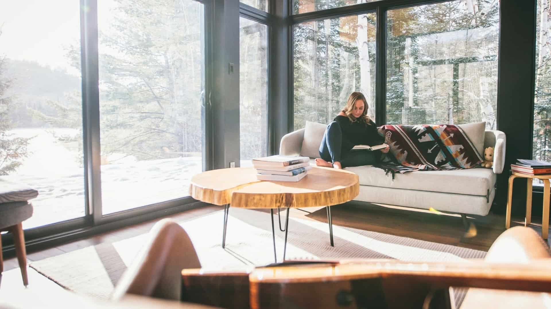 Woman working on laptop in a bright, modern living room with large glass windows.