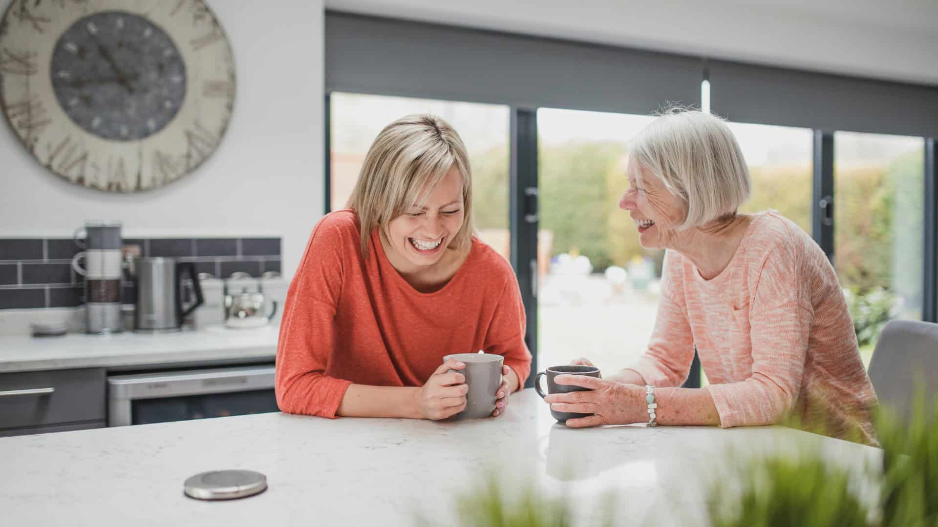Two women smiling and using smartphones in a modern kitchen.